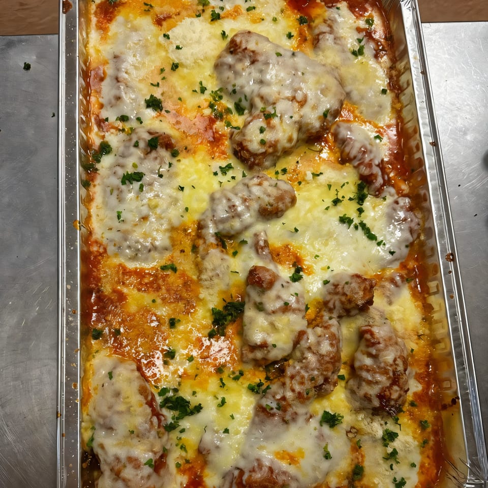A large aluminum tray filled with baked chicken parmesan, topped with melted cheese and parsley.
