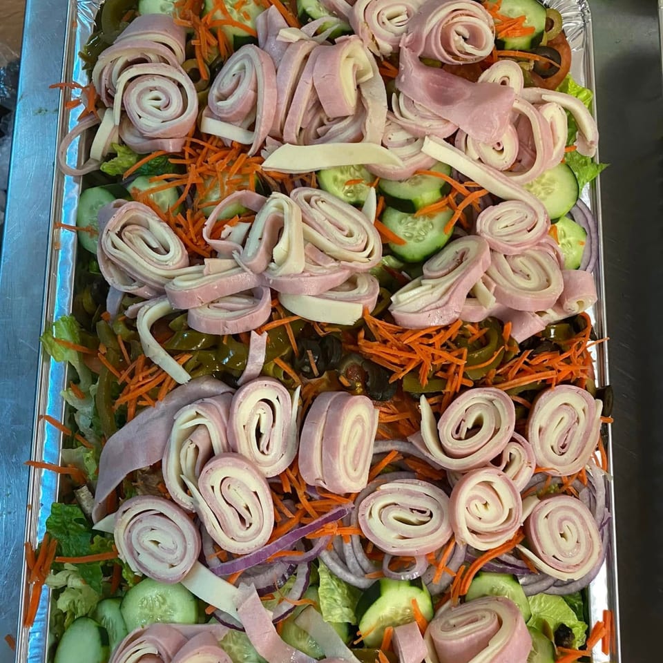 A large catering tray filled with a fresh salad topped with rolled ham and cheese, vegetables.