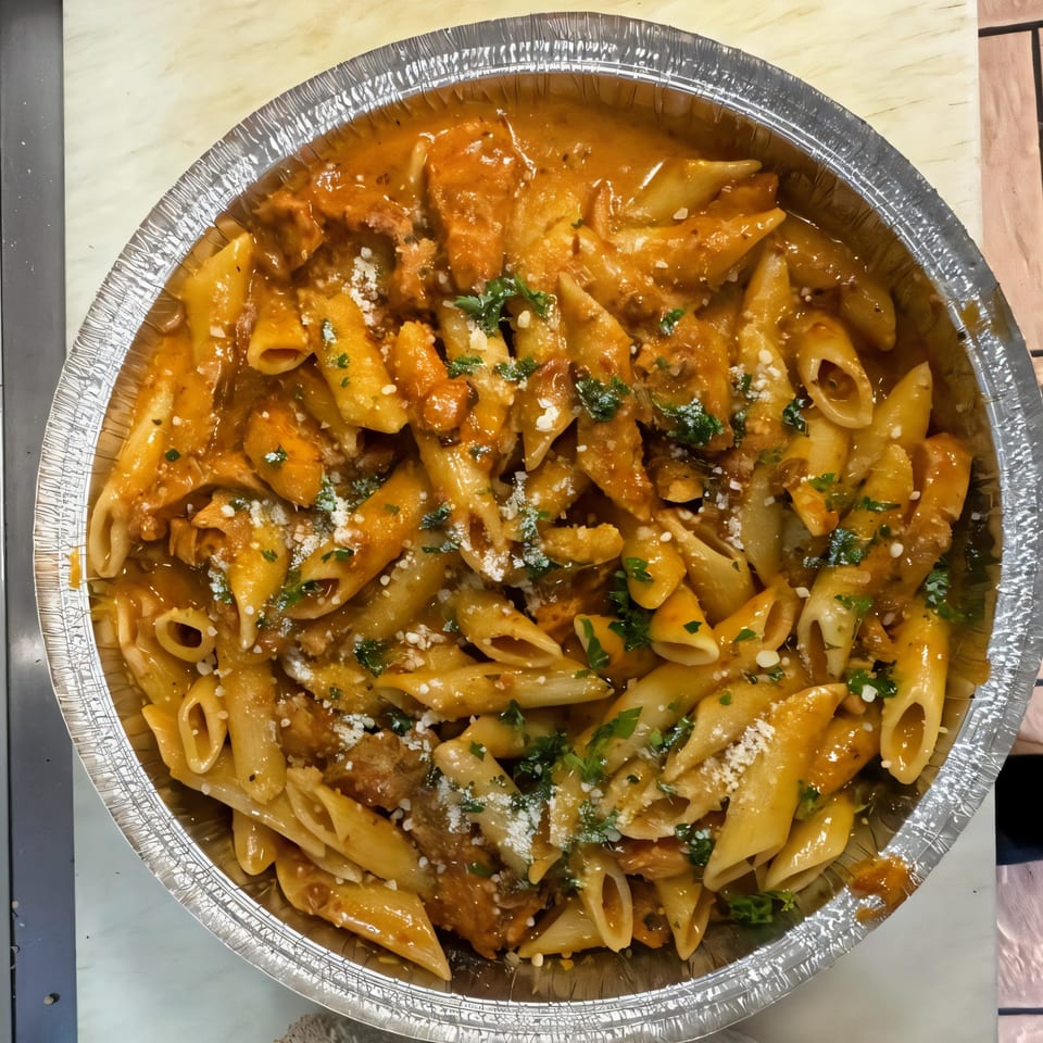 Penne pasta with chicken in a creamy tomato sauce, topped with parmesan and parsley, in a foil pan.
